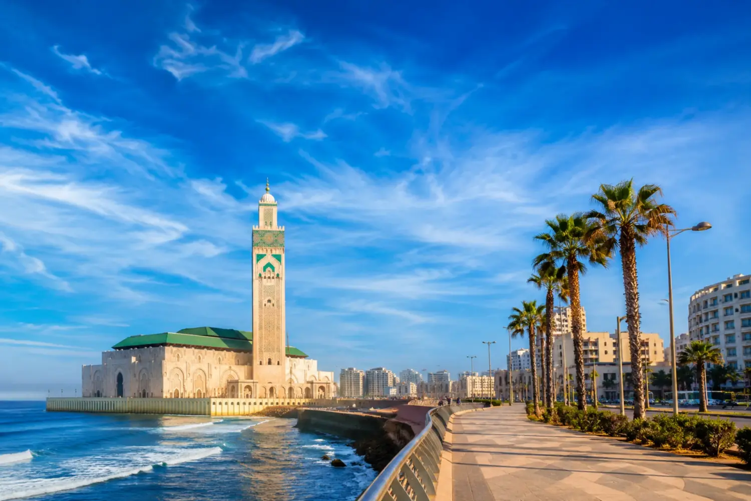 Hassan II Mosque in Casablanca overlooking the Atlantic Ocean, with palm-lined waterfront and clear blue sky.
