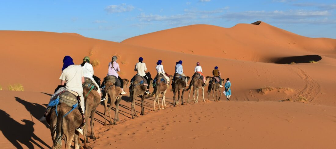 Best Morocco Desert Tours in 2025 – Complete Guide for USA & Europe Travelers Camel ride in the Merzouga Sahara desert, Morocco – travelers crossing Erg Chebbi dunes at sunset