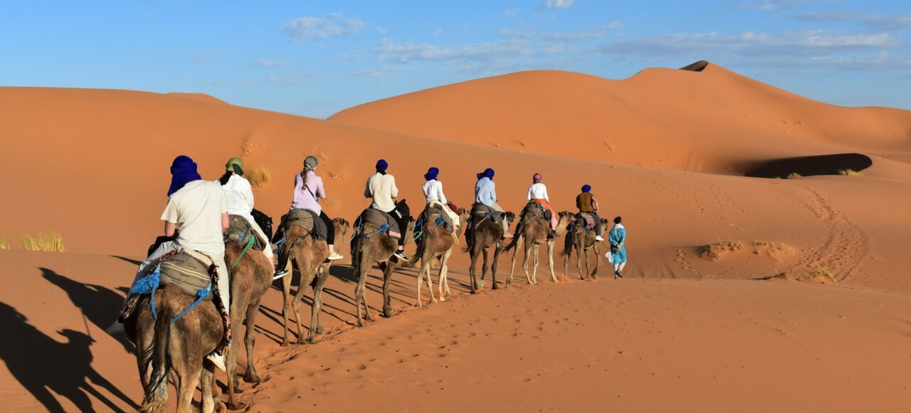 Best Morocco Desert Tours in 2025 – Complete Guide for USA & Europe Travelers Camel ride in the Merzouga Sahara desert, Morocco – travelers crossing Erg Chebbi dunes at sunset