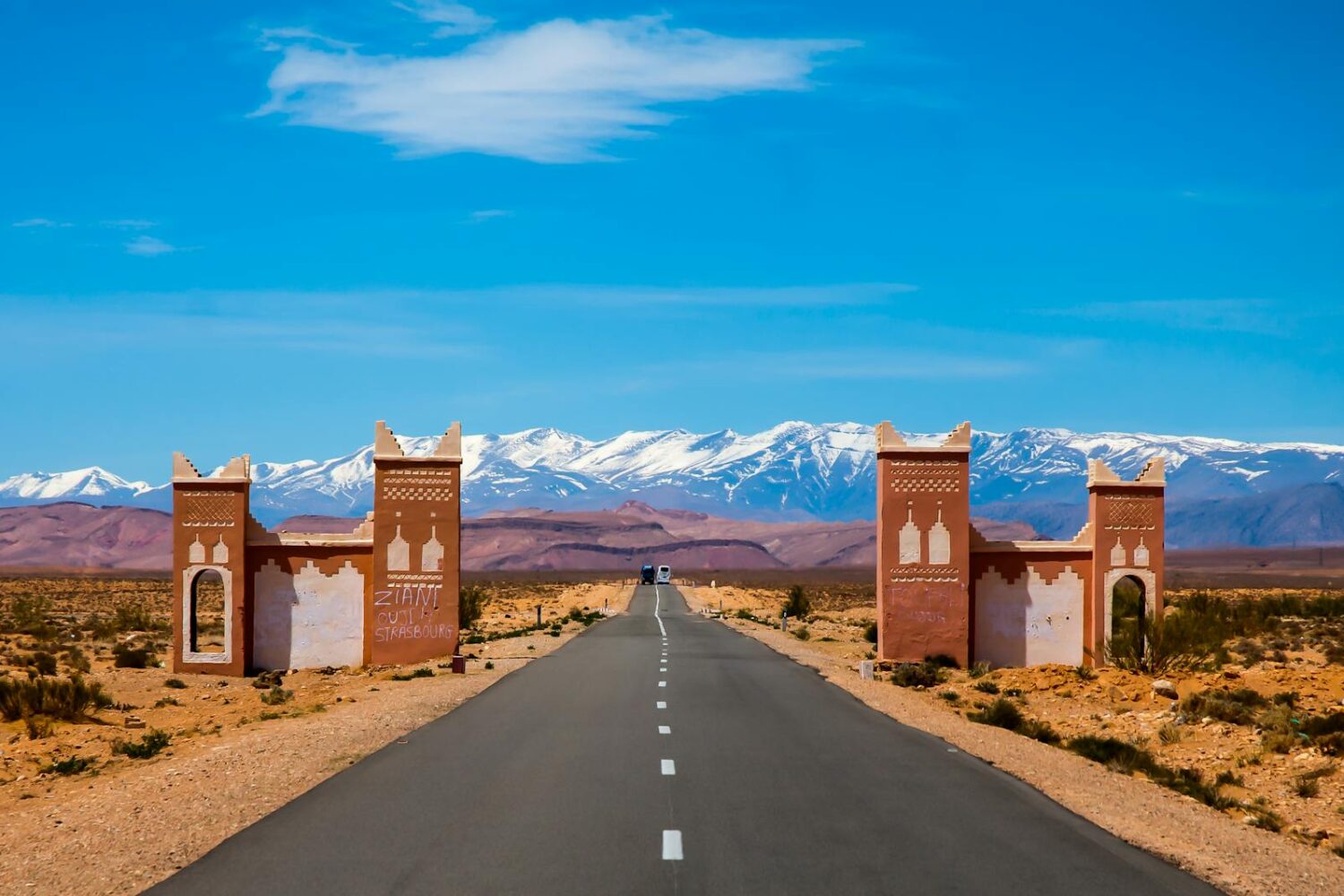 Desert road with kasbah-style gate and Atlas Mountains, part of the 3 Days / 2 Nights Marrakech to Fes via Merzouga Desert Tour