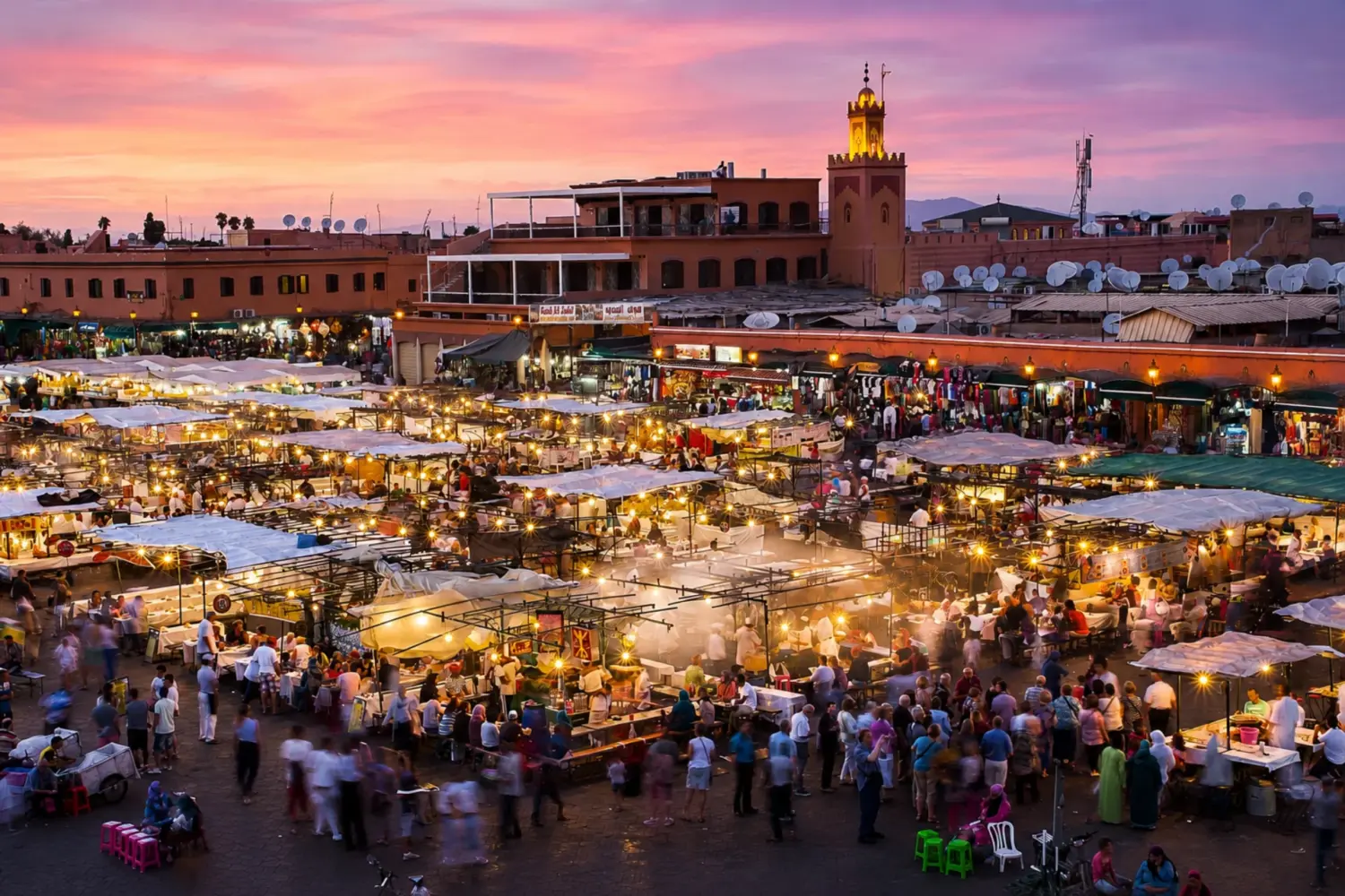 Panoramic view of Marrakech's Jemaa el-Fna Square at sunset with colorful market stalls, crowds, and the illuminated Argana Mosque.