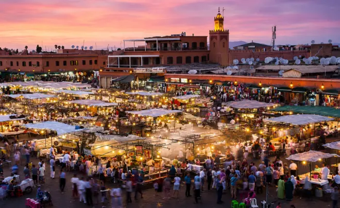 Panoramic view of Marrakech's Jemaa el-Fna Square at sunset with colorful market stalls, crowds, and the illuminated Argana Mosque.