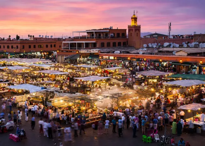 Panoramic view of Marrakech's Jemaa el-Fna Square at sunset with colorful market stalls, crowds, and the illuminated Argana Mosque.