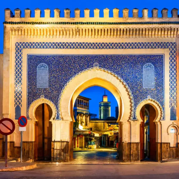 Illuminated Bab Bou Jeloud gate in Fes featuring intricate blue zellige tilework and traditional Moroccan architecture at night during 3 Day Tour from Fes to Marrakech via Chefchaouen