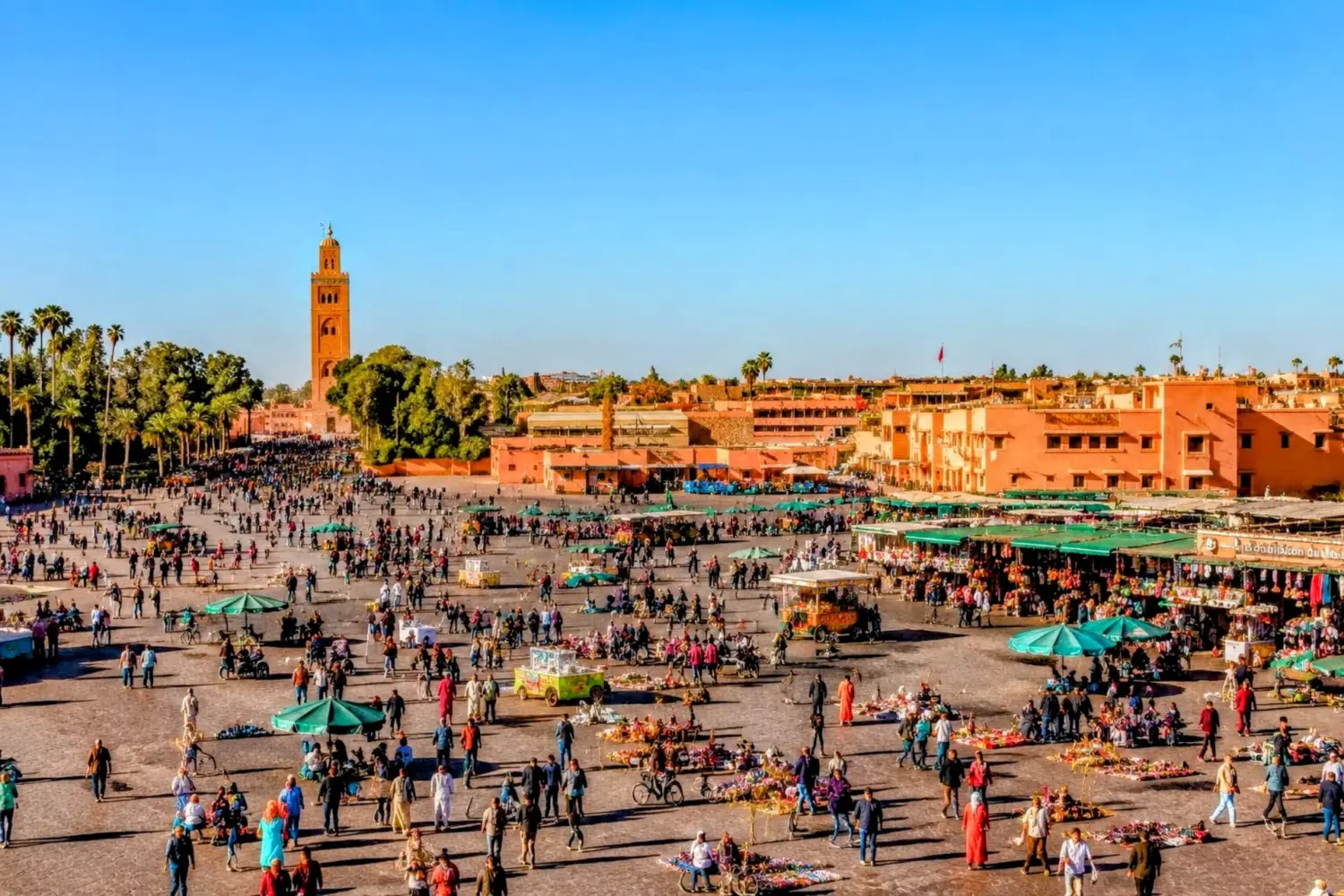 Jemaa el-Fnaa Square in Marrakech on a sunny day with a clear blue sky, bustling market stalls, local vendors, visitors, and the Koutoubia Mosque in the background.
