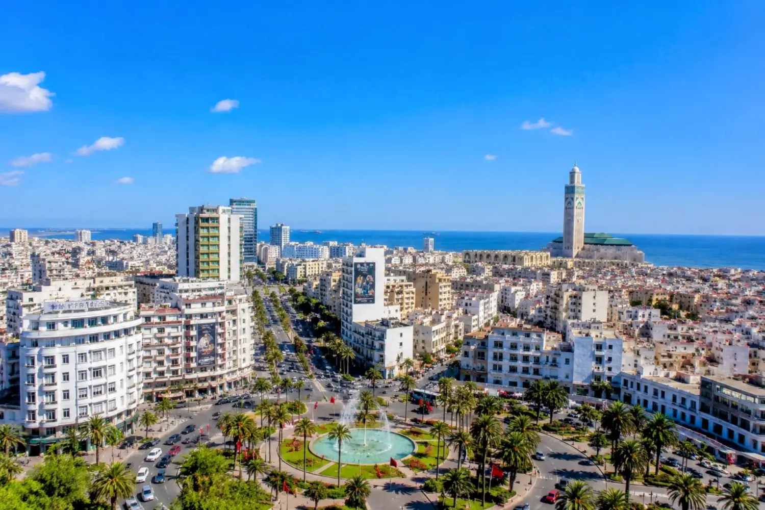 Panoramic view of Casablanca city with Hassan II Mosque, palm-lined streets, modern buildings, and a clear blue sky during 5 Day Chefchaouen Fes and Sahara Tour from Casablanca.