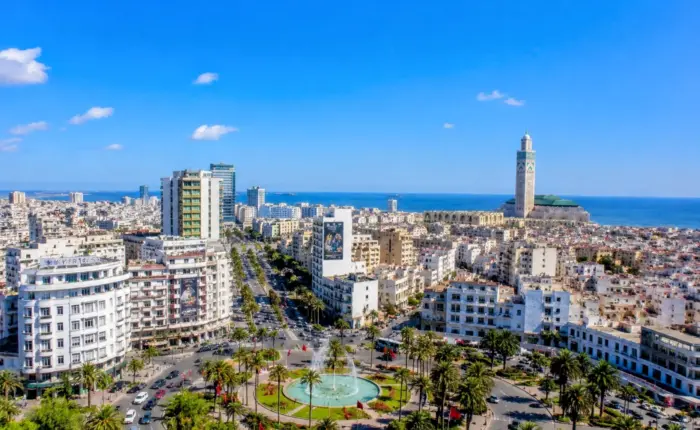 Panoramic view of Casablanca city with Hassan II Mosque, palm-lined streets, modern buildings, and a clear blue sky during 5 Day Chefchaouen Fes and Sahara Tour from Casablanca.